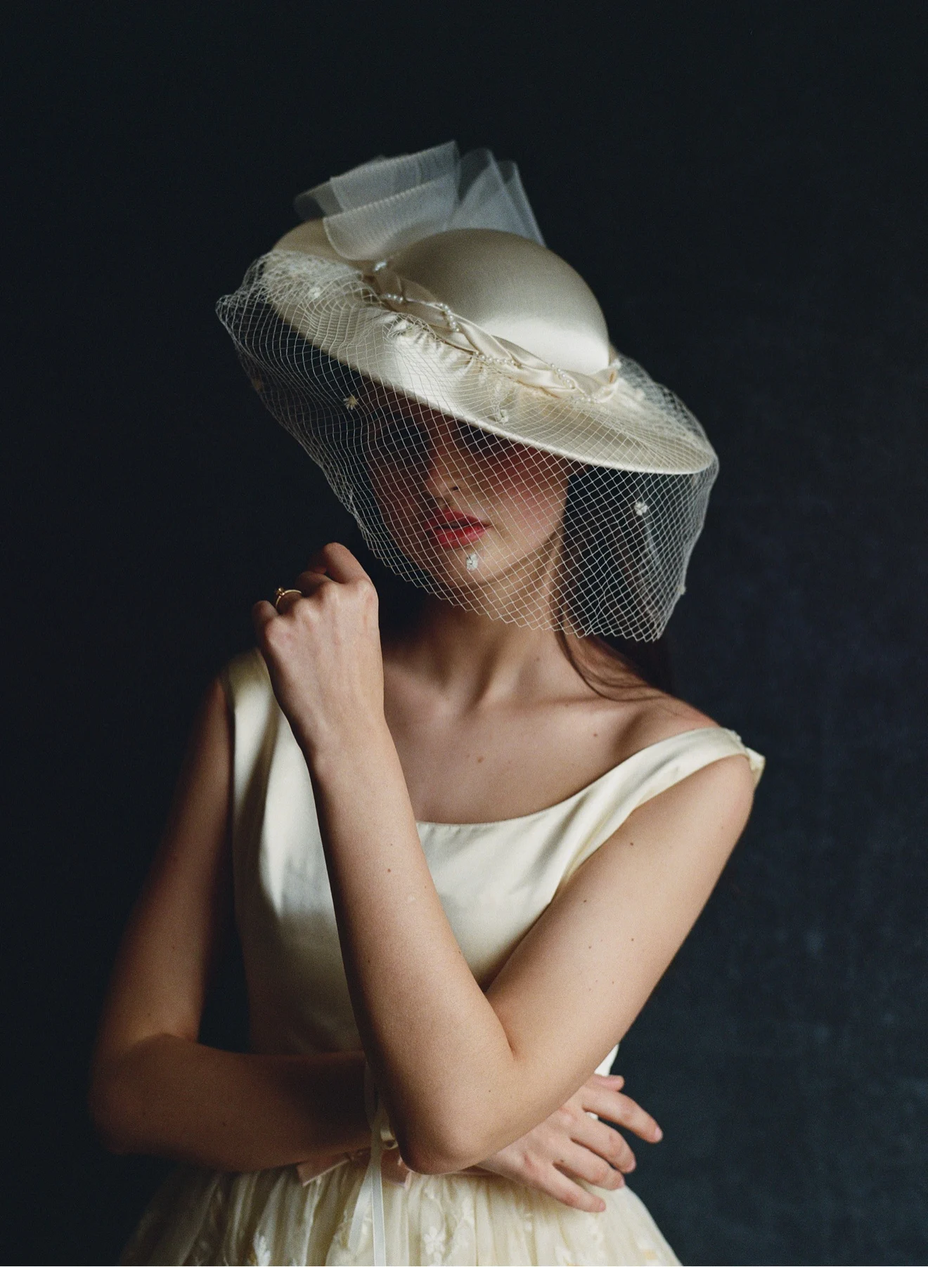 Bride in profile wearing an ivory couture hat with a delicate net veil, against a dark studio backdrop