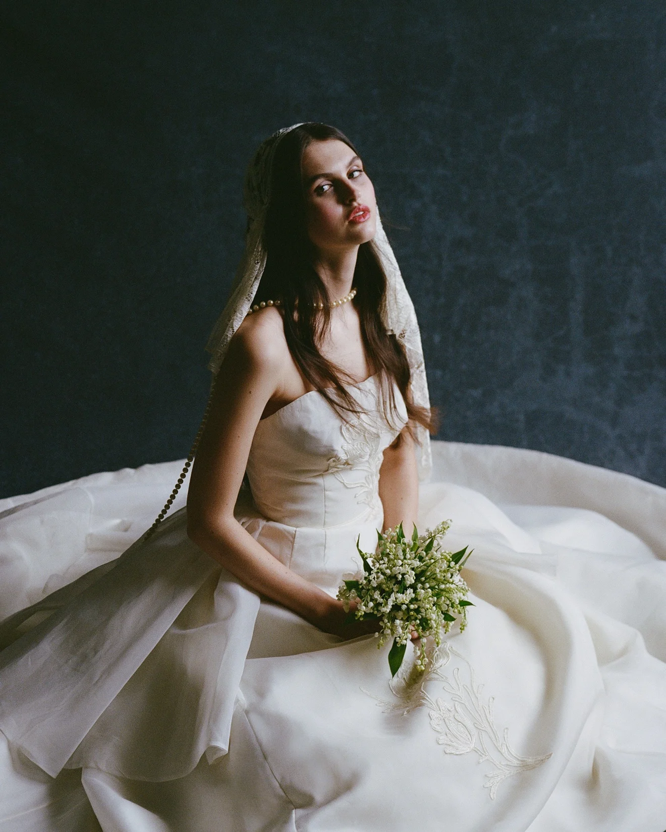 Seated bride in an ivory gown and lace mantilla veil, holding a lily-of-the-valley bouquet against a dark studio backdrop