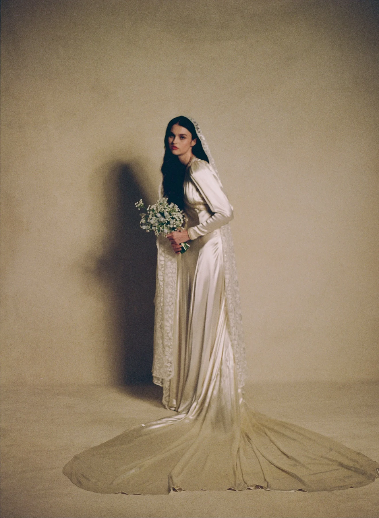 Bride in a full-length vintage satin gown with a lace-edged veil and sweeping train, holding a small white bouquet against a warm studio backdrop
